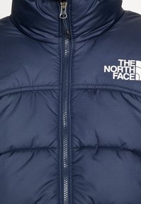 Mörkblå vadderad jacka med hög krage, framdragkedja och vit logotyp på vänster bröst. Texturerat tyg med quiltade sektioner.