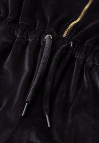 Zwarte fluweelstof met een elastische tailleband, trekkoorden door metalen oogjes en een zichtbare gouden ritssluiting aan de rechterbovenkant.