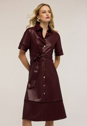 Kobieta ubrana w bordową skórzaną sukienkę z krótkimi rękawami, zapinaną z przodu na guziki, z paskiem przewiązanym w talii oraz dużymi złotymi kolczykami kołami na neutralnym tle.