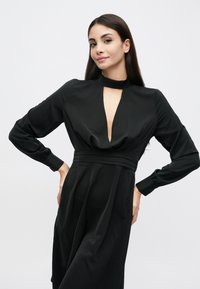 Rochie neagră cu decolteu adânc în V și guler înalt. Mâneci lungi cu mansete strânse. Material neted și fluid cu detalii strânse în talie.