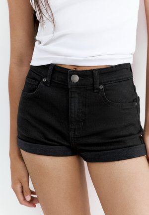 Femme portant un short en denim noir taille haute avec des revers roulés et un débardeur court blanc, debout devant un fond uni.