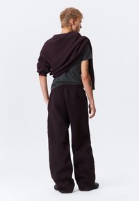 Bordeauxfarbenes Sweatshirt über den Schultern, graues T-Shirt und weit geschnittene dunkle Hose mit floralen Stickereien und Rückentaschen.