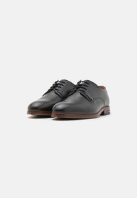 Chaussures en cuir noir à lacets avec un bout arrondi, semelle texturée marron et détails subtils près des œillets. Fini lisse sur l'ensemble.