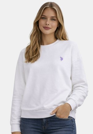Junge Frau mit langen blonden Haaren, trägt ein weißes Sweatshirt mit kleinem lila Logo, Hand in der Tasche, steht vor einem einfarbigen Hintergrund.