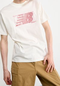 Camiseta crema hecha de una mezcla de algodón, con un diseño gráfico geométrico rojo de la ‘N’ con círculos y líneas impresos en el frente.
