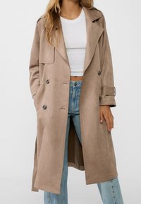 Beige dlouhý trenčkot vyrobený z hladkého semiše, s dvojitým zapínáním, velkými kapsami a ohrnutými rukávy. Stylizován přes bílý top a džíny.