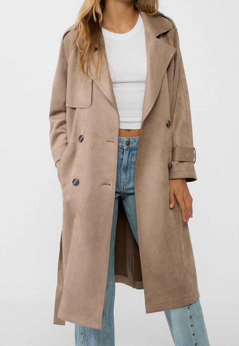 Beige dlouhý trenčkot vyrobený z hladkého semiše, s dvojitým zapínáním, velkými kapsami a ohrnutými rukávy. Stylizován přes bílý top a džíny.