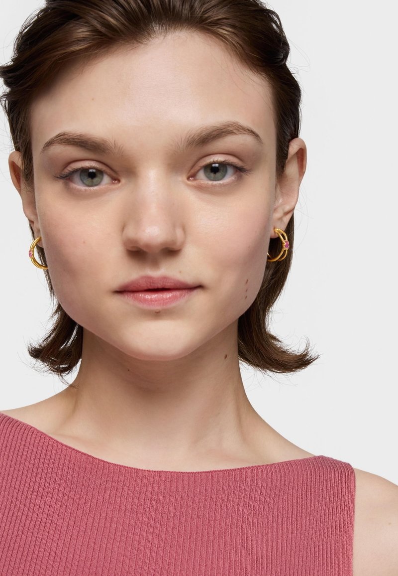 Orecchini dorati rotondi con un accento rosa indossati da un modello. Il modello ha un taglio di capelli corto e texturizzato ed è vestito con una maglietta rosa a coste.