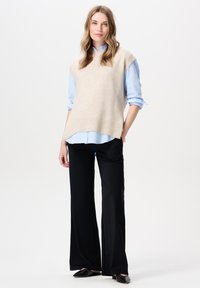 Beige gebreide vest over een lichtblauw overhemd, gecombineerd met zwarte wijde broekspijpen. Zachte textuur, ontspannen pasvorm, ribgebreide zoom op het vest.