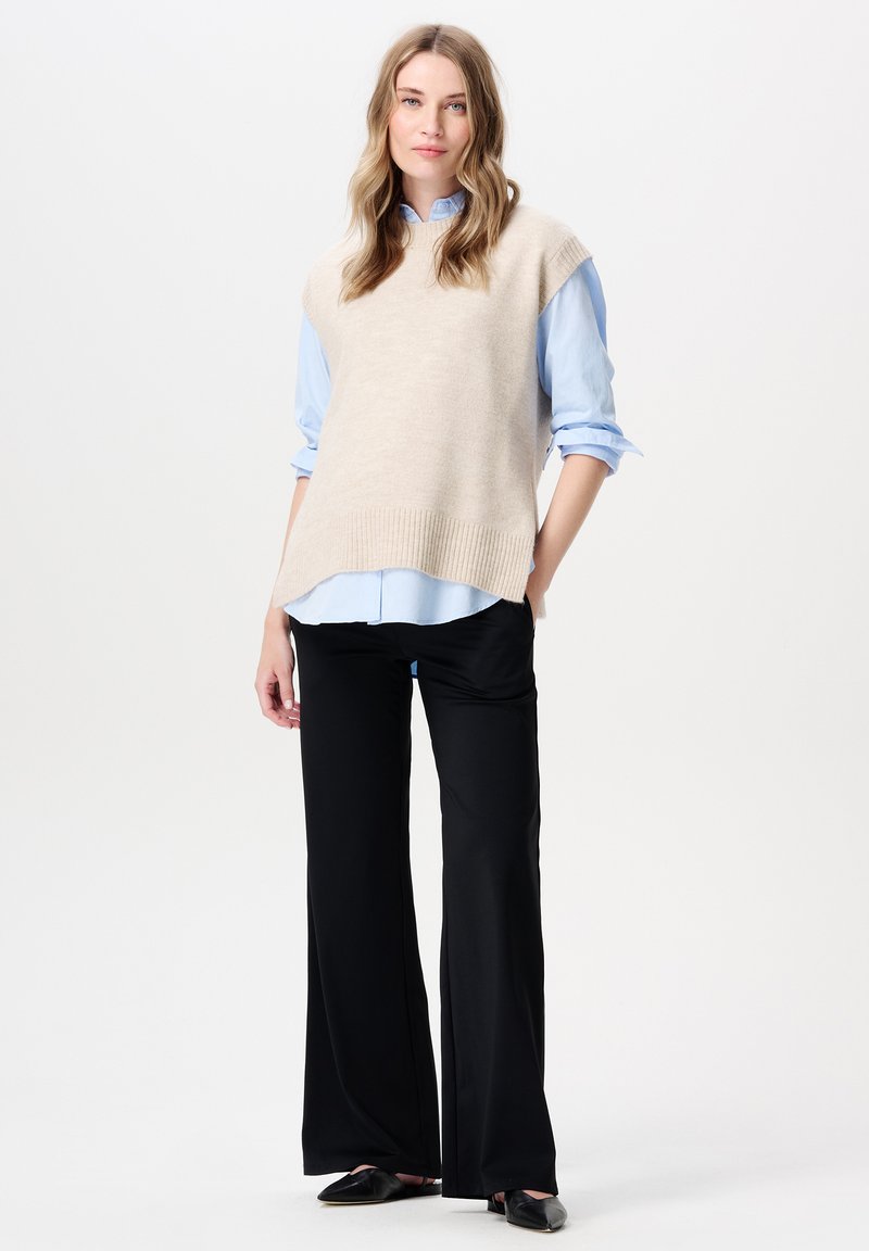 Beige gebreide vest over een lichtblauw overhemd, gecombineerd met zwarte wijde broekspijpen. Zachte textuur, ontspannen pasvorm, ribgebreide zoom op het vest.