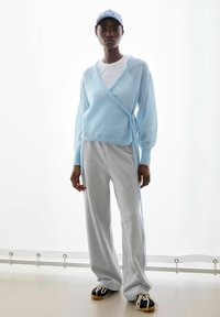 Lichtblauwe wikkeltrui gecombineerd met grijze sweatpants met brede pijpen. De outfit heeft zachte materialen, een relaxte pasvorm en contrasterende strepen aan de zijkant.