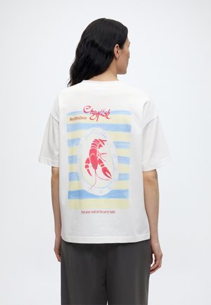 Άτομο με μακριά σκουρόχρωμα μαλλιά φοράει λευκό μπλουζάκι που δείχνει έναν κόκκινο αστακό σε ριγέ μπλε και κίτρινο φόντο με το κείμενο "English" και "find your seat" στην πλάτη.