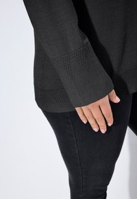 Person in einem dunkel strukturierten langärmeligen Pullover und schwarzen, enganliegenden Hosen, mit der Hand sanft an ihrer Seite vor einem hellen Hintergrund.