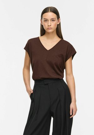 Jeune femme aux cheveux bruns lisses portant un top marron à col en V et un pantalon noir taille haute, une main dans la poche.