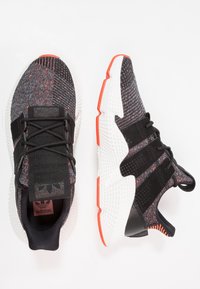 Svarta och grå sportskor med ett texturerat stickat överdel, vit mellansula och orange accenter. Har en snörningsdesign och vadderad krage.