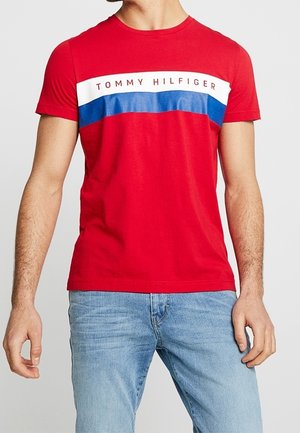 Man wearing a red Tommy Hilfiger t-shirt with white and blue horizontal stripes, paired with light blue jeans.