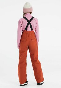 Orange Skihosen mit schwarzen verstellbaren Trägern, kombiniert mit einem langärmligen pinken Oberteil und einer hellrosa Strickmütze, von hinten getragen.