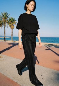 Young woman dressed in black walking on a seaside promenade with palm trees and ocean in the background under clear sky.