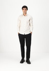 Beige langermet skjorte med en tettsittende design, knapp foran, og en glatt tekstur, parret med svarte tapered bukser og svarte sko.