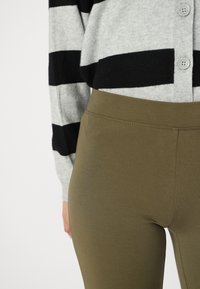 Olivno zeleni leggingi, izdelani iz mehkega materiala, z gladko teksturo in visokim pasom. V kombinaciji s sivo-črnimi črtastimi puloverjem.