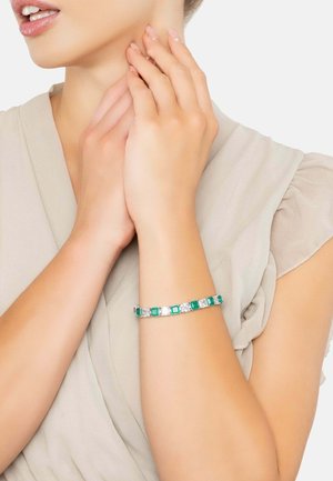 Silver bracelet with alternating square green and clear gemstones. Smooth surface and geometric design. Worn on a bare wrist.