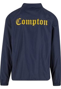 Jachetă impermeabilă de marină, cu text galben "Compton" pe spate. Prezintă un guler și manșete elastice, realizată dintr-un material ușor.