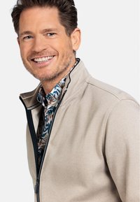 Beige zip-up jack met een gestructureerde stof en een contrasterende marineblauwe rits, gelaagd over een bloemrijke blouse met groene en oranje accenten.