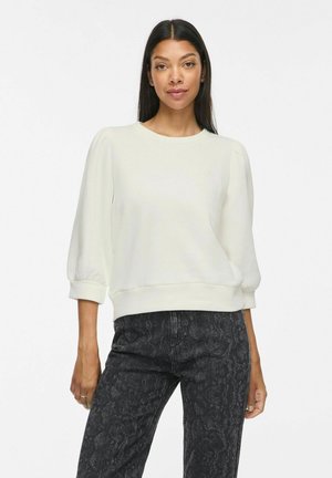 Femme aux longs cheveux foncés portant un chemisier blanc à manches bouffantes et un jean noir à motifs taille haute, debout devant un fond uni.