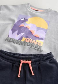 Pull gris avec un graphique de dinosaure en violet, des accents roses et le texte "Aventures Dino". Short noir avec cordon de serrage doté d'une taille élastique.
