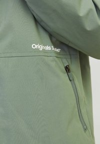 Zielony materiał kurtki z białym detalem logotypu "Originals Studio" oraz pionową kieszonką na zamek po boku.