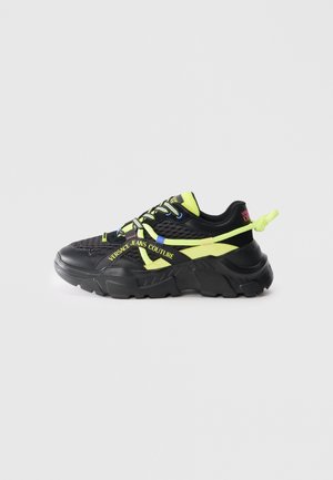 Black sneakers with mesh and synthetic upper, yellow accents, and bold branding. Chunky sole offers a textured, sporty design.