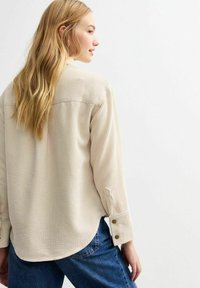 Beige corduroy shirt with long sleeves, featuring a back yoke, buttoned cuffs, and a slightly curved hem. Worn with blue denim pants.