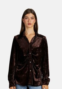 Camicia button-up in velluto marrone con colletto, caratterizzata da un design drappeggiato e maniche lunghe. Include bottoni decorativi sulla parte frontale.