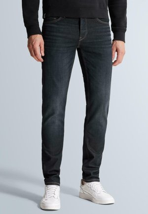 Muž s tmavými slim-fit džínsami, čiernou mikinou a bielymi teniskami stojaci pred jednoduchým svetlým pozadím.