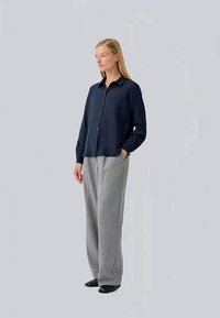 Woman standing, wearing a navy buttoned shirt, gray wide-leg pants, and black shoes, with one hand in pocket against a plain background.