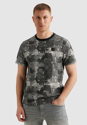 Junger Mann mit kurzen Haaren, der ein schwarz-grau gemustertes T-Shirt mit kurzen Ärmeln und hellgraue Jeans trägt, Hand in der Tasche.