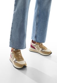 Gambe che indossano jeans in denim azzurri chiari e sneakers beige, grigie, bianche e bordeaux, in piedi su una superficie bianca.