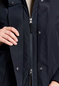 Marinblå vattentät jacka med tvåvägsdragkedja, tryckknappar och sidofickor. Har en slät yta och matt finish.