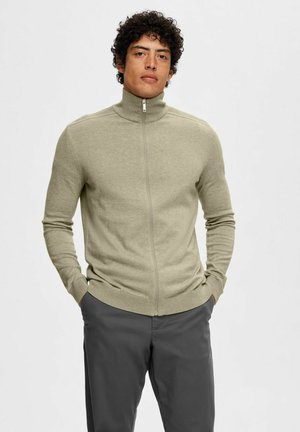 Sudadera de cremallera verde claro con cuello alto, diseño ajustado, puños y dobladillo acanalados. Lleva puesta sobre pantalones gris oscuro. Textura suave.