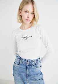 Valkoinen pitkähihainen paita, jossa on musta "Pepe Jeans London" -logo, yhdistettynä korkeavyötäröisiin vaaleansinisiin farkkusiin, joissa on joustava vyötärö ja taskut.
