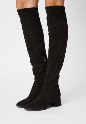 Bottes montantes en daim noir avec talons carrés et fermetures éclair latérales, portées sur des jambes nues sur fond blanc.