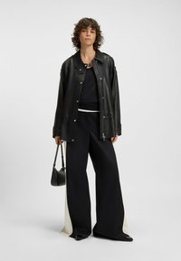 Black leather jacket with snap closures, paired with loose black trousers featuring white side panels and glossy black shoes. Small black handbag.