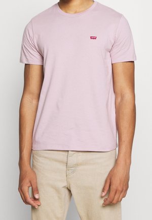 Mężczyzna w jasnoróżowej koszulce Levi's i beżowych spodniach, stojący ze swobodnie opuszczonymi ramionami na tle jednolitego koloru.