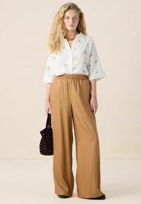 Witte shirt met schelpenpatroon, losse pasvorm, wijdvallende bruin satijnen broek, houdt een kleine zwarte tas met rond uitsnede ontwerp vast.