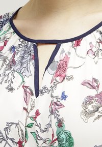 Blommig blus med flerfärgad botanisk tryck, lättviktsmaterial, marinblå kant runt halsen, med ett nyckelhålsdetalj fram.