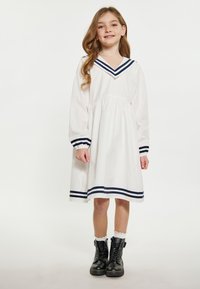 Weißes Kleid mit V-Ausschnitt, marineblau gestreiftem Kragen und Manschetten sowie einem gerafften Taillenteil. Getragen mit schwarzen Stiefeln und weißen Knöchelsocken.
