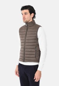 Gilet imbottito in nylon marrone con colletto alto, chiusura a zip e tasche laterali. Indossato sopra una camicia bianca a maniche lunghe e pantaloni blu navy.