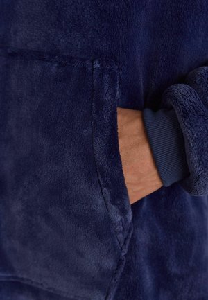 Marinblå plyschhoodie med mjuk textur, med en hand i fickan och ribbade ärmslut. Sömmar är synliga på tyget.