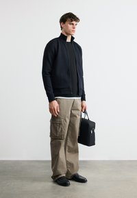 Άνδρας που φοράει μαύρο μπουφάν με φερμουάρ, μαύρο πουκάμισο, μπεζ παντελόνι cargo, μαύρα παπούτσια, κρατώντας μια μαύρη τσάντα γραφείου σε απλό λευκό φόντο.