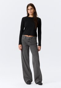 Schwarzes Crop-Top mit langen Ärmeln, kombiniert mit locker sitzenden grauen Jeans. Die Jeans haben ein verblasstes Finish und Vordertaschen. Schwarze Schuhe runden das Outfit ab.